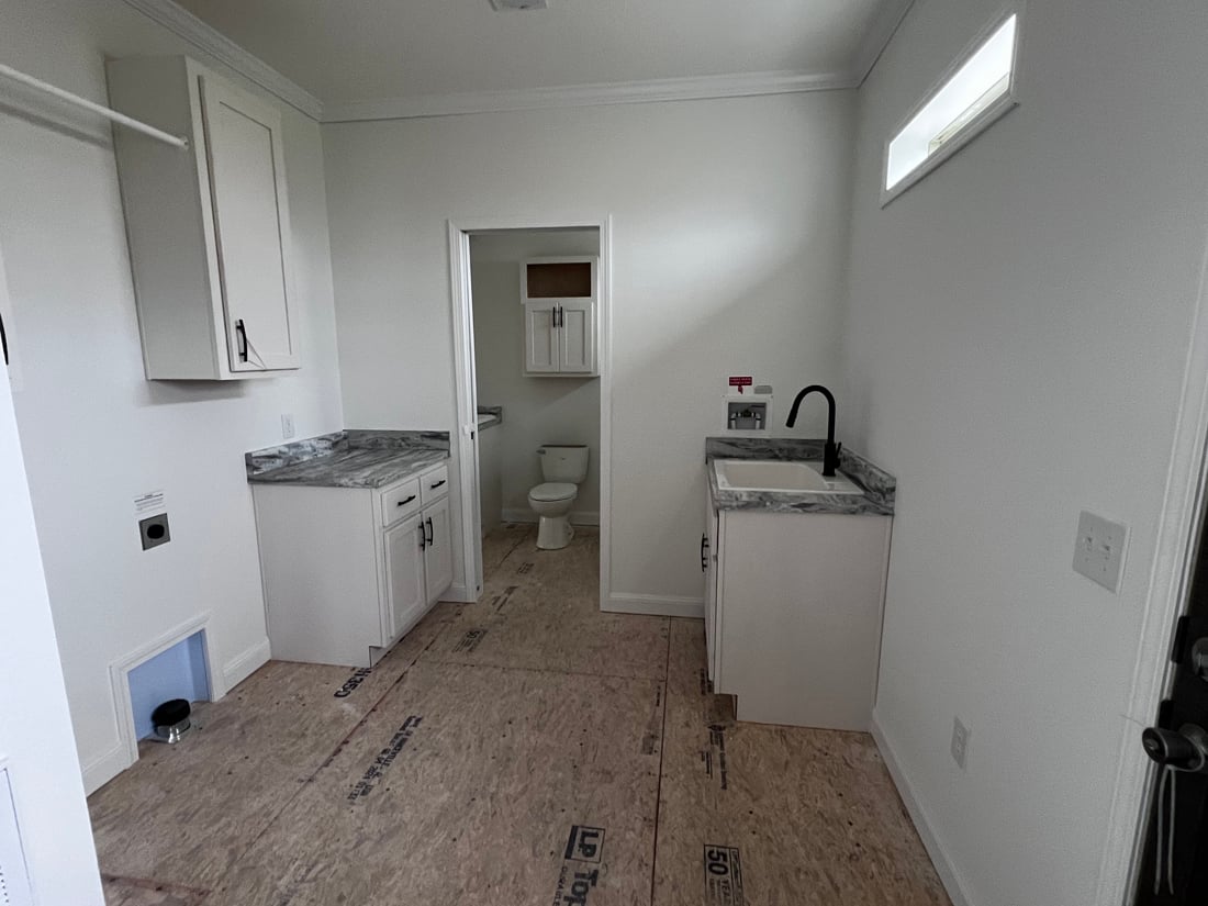 A small laundry room with white walls, unfinished wood flooring, and marble countertops. It features cabinets, a black faucet, and a visible toilet in an adjacent room. The space feels clean and minimalistic.