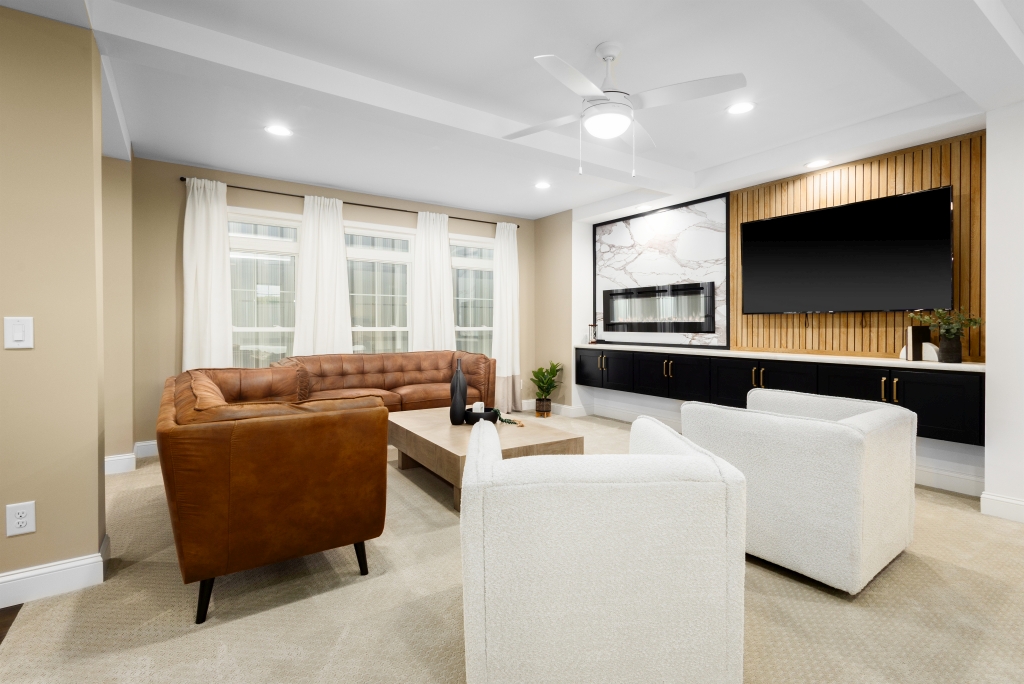 Modern living room with beige carpet, brown leather sectional, and white armchairs. A wall-mounted TV and fireplace sit above a dark cabinet.
