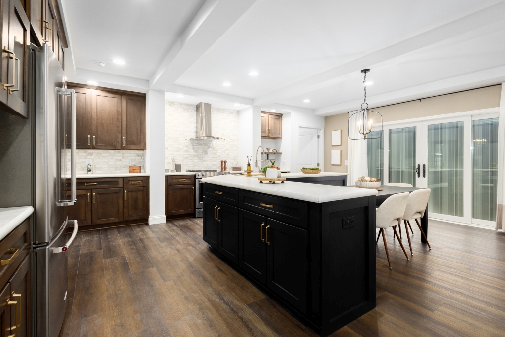 A modern kitchen with dark wood cabinets, central island with white countertop, pendant light, and dining table. Large patio doors provide natural light.