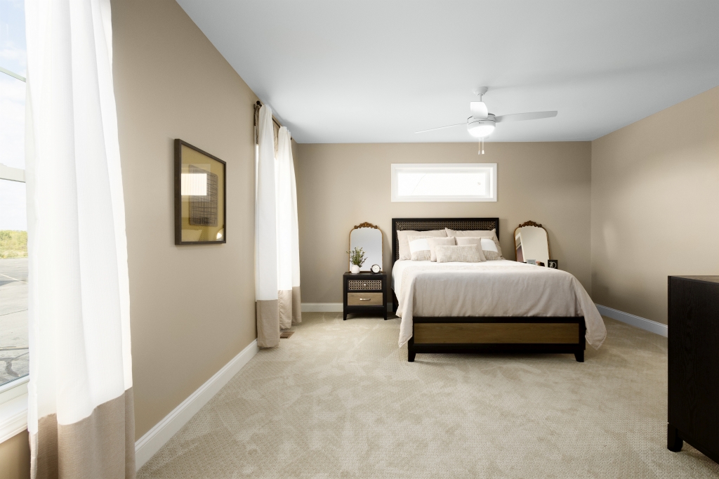 A cozy bedroom with beige walls and carpet, featuring a neatly made bed with brown frame, matching nightstands, table lamps, and soft natural light.
