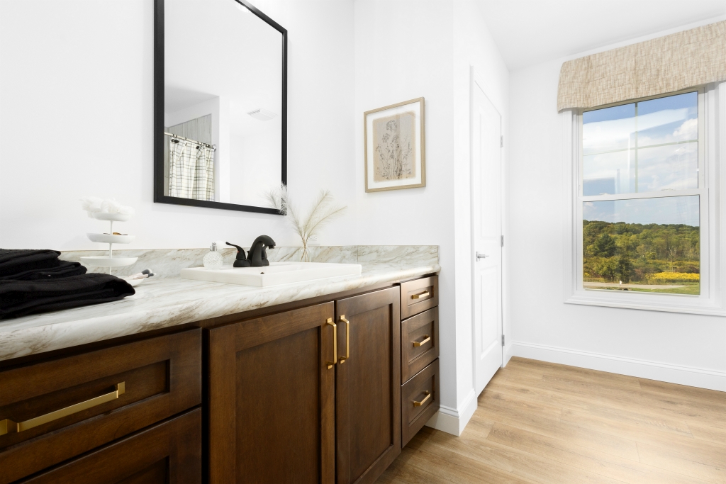 A bright bathroom features a dark wood vanity with gold handles, a marble countertop with a black faucet. A large mirror, window with scenic view, and wood floor add elegance.
