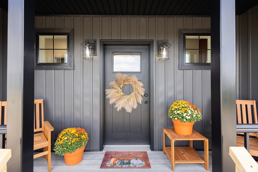 Cozy porch with dark door, wreath, and lanterns. Bright orange and yellow potted flowers flank entrance. Wooden benches add warmth and comfort.
