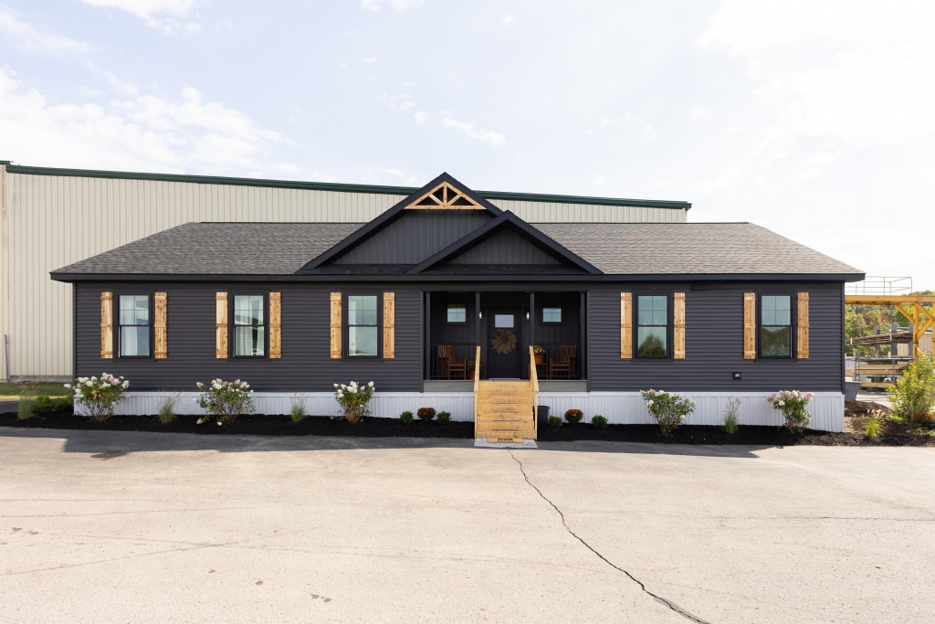 A modern, dark gray, single-story house with wooden shutters and a small front porch, featuring a gabled roof and surrounded by minimal landscaping.