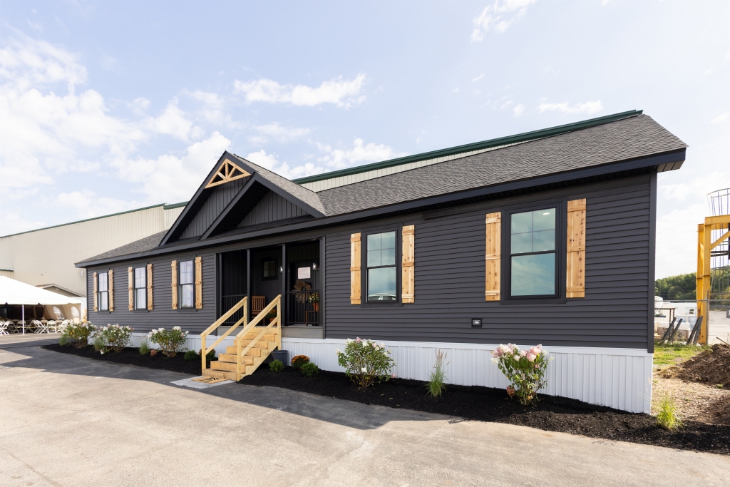 A modern, single-story manufactured home with dark gray siding, wooden shutters, and a welcoming front porch. It's surrounded by neat landscaping under a bright, partly cloudy sky.