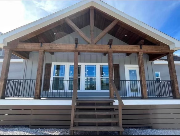 A modern house exterior with a wooden porch and gabled roof. The house features large glass doors, wooden beams, and a staircase leading to the entrance.