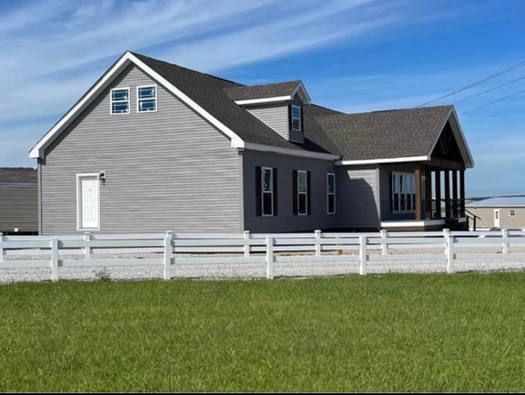 A gray house with a dark roof sits behind a white fence, surrounded by green grass. The sky is clear and blue, creating a calm, peaceful setting.