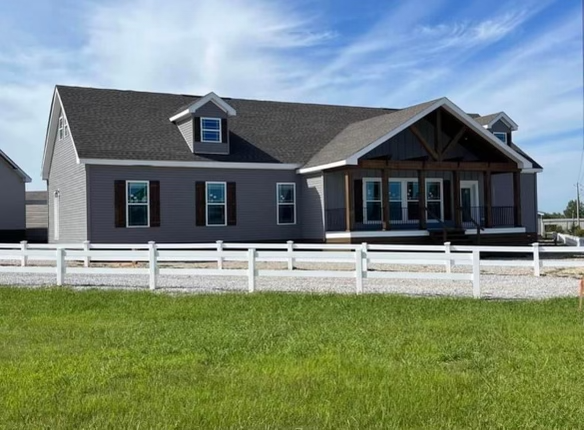 A modern gray house with a front porch and large windows, surrounded by a white fence. It sits on a green lawn under a clear, blue sky.