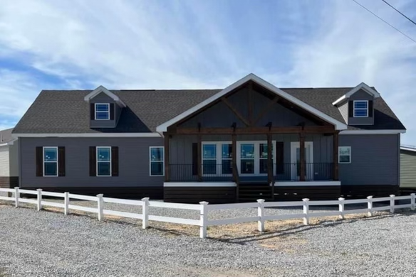 Alt text: A modern, single-story house with a dark gray facade and a pitched roof. It features a central covered porch, white trim, and a neatly gravelled yard, enclosed by a white picket fence under a clear blue sky.