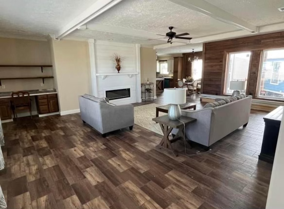 Open living room with wooden floors, two gray sofas facing a white fireplace. A ceiling fan hangs above. Large windows let in natural light. Cozy and inviting atmosphere.