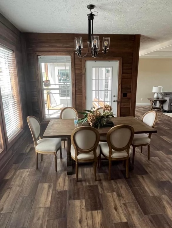 Warm dining room with wood-paneled walls and flooring, featuring a wooden table, six cushioned chairs, a central floral arrangement, and a windowed door.
