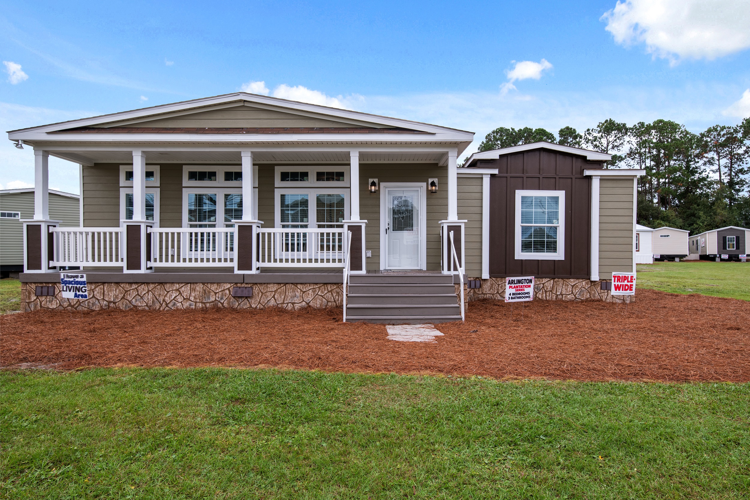 A beige manufactured home with large windows and a front porch sits under a blue sky. Signs indicate "Triple Wide" and "Arlington Living."