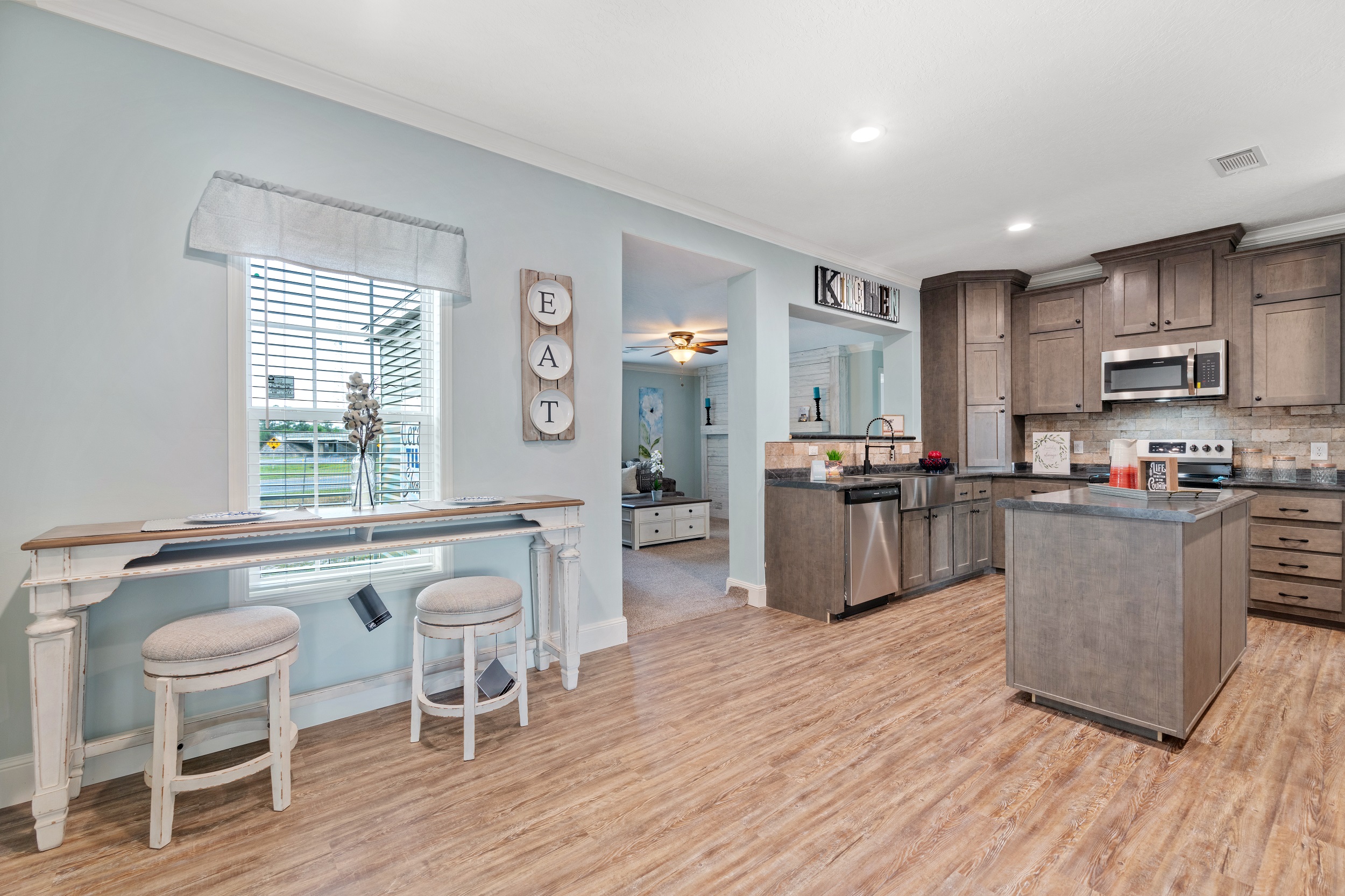 Modern kitchen with light blue walls and wood flooring. Features dark wood cabinets, a central island, stainless steel appliances, and decor reading “EAT” and “KITCHEN.” Cozy, inviting atmosphere.