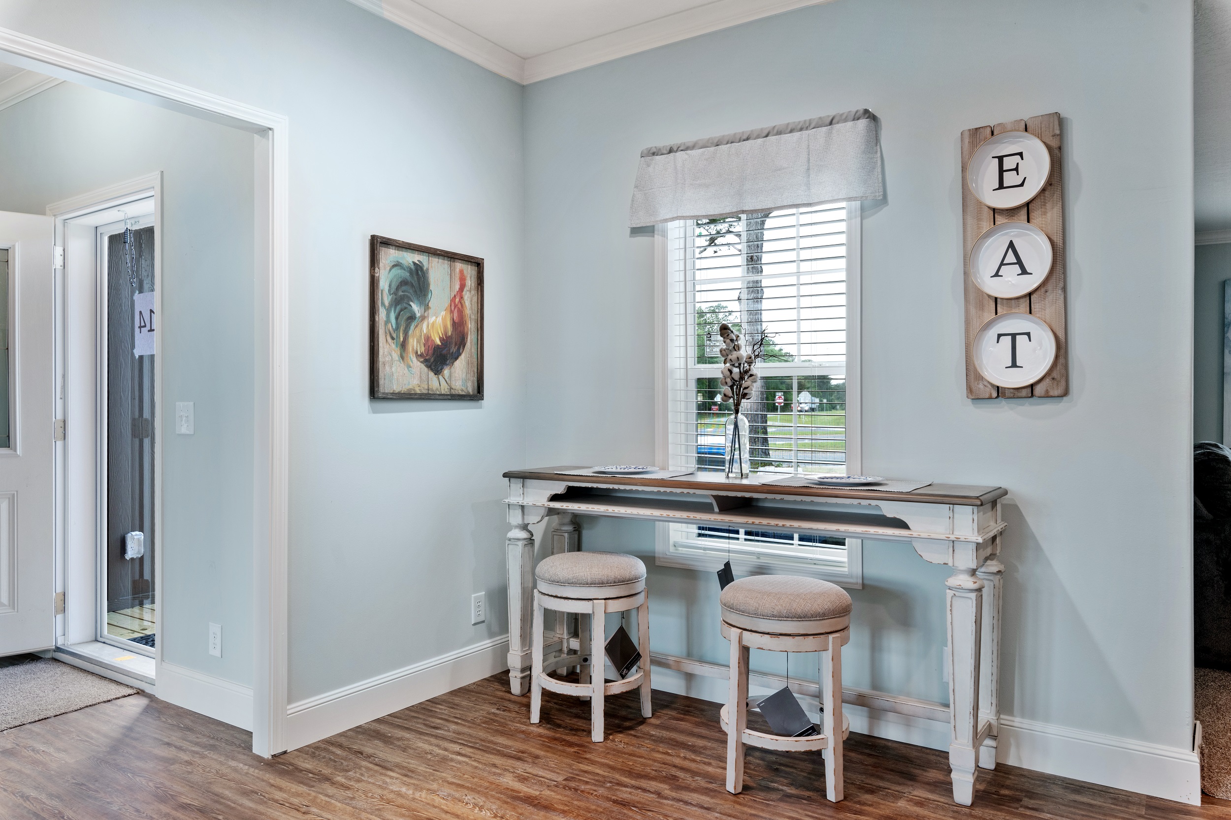 Stylish kitchen nook with light blue walls, white desk, two round stools, and window. Rooster art and "EAT" sign add charm. Homey and inviting.