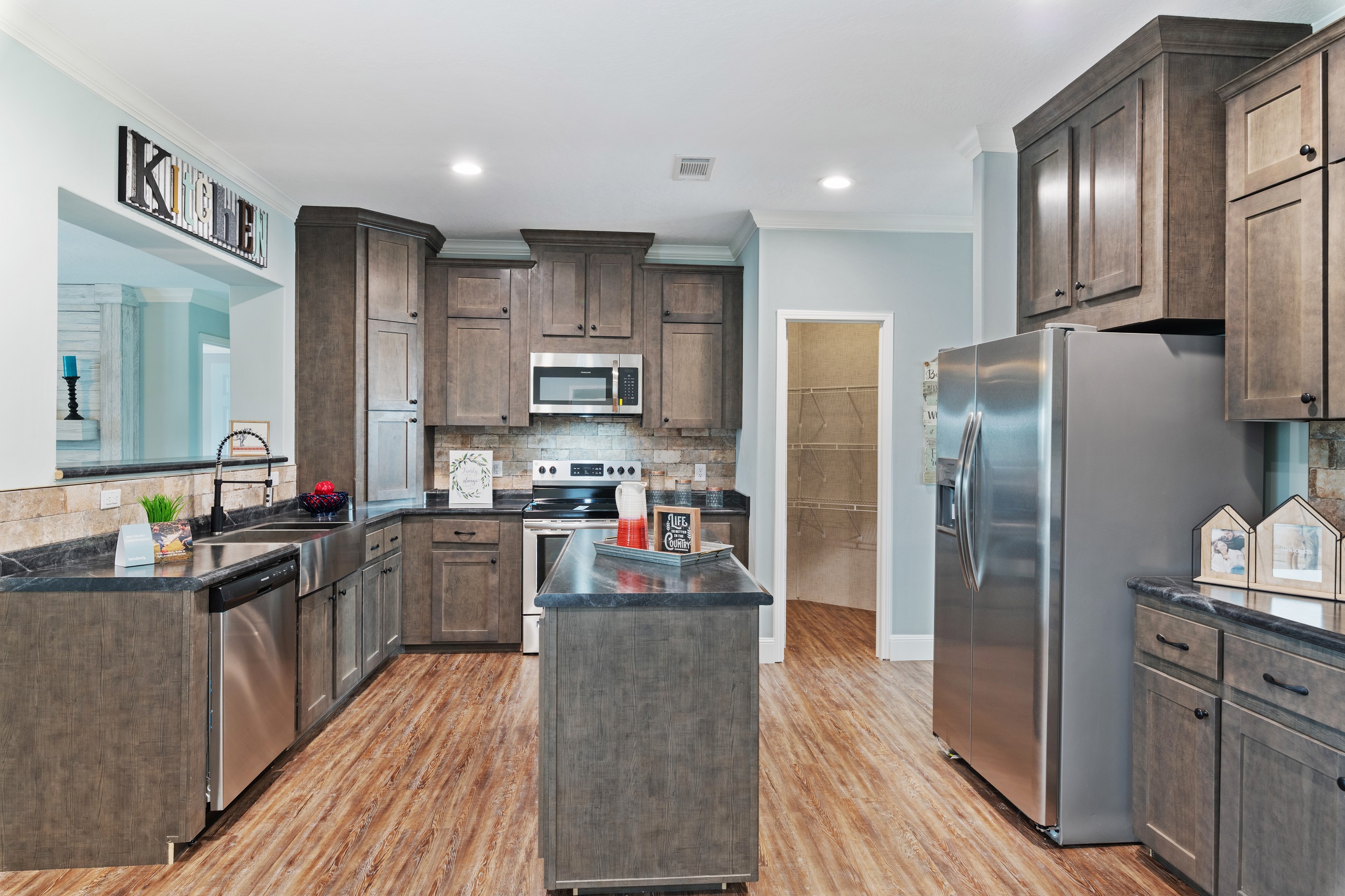 Modern kitchen with dark wooden cabinets, stainless steel appliances, and a large fridge. There's an island in the center and hardwood flooring.