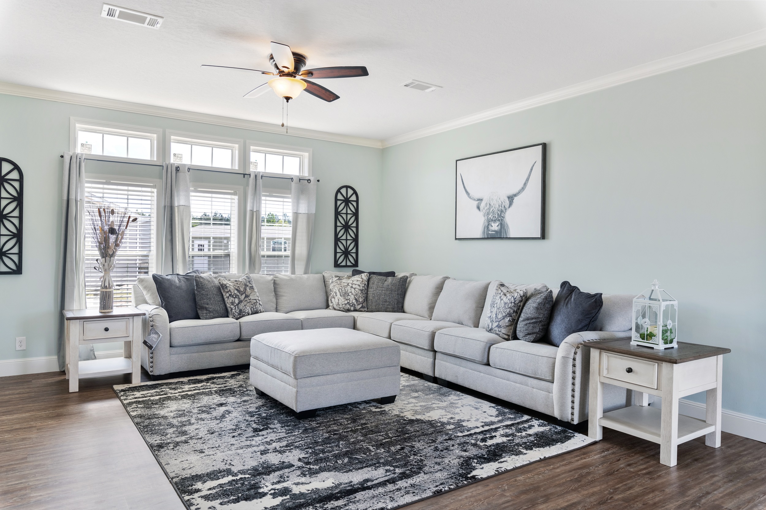 A cozy living room with a large gray sectional sofa, abstract rug, and bull artwork. Soft lighting and pastel walls offer a calm, inviting atmosphere.