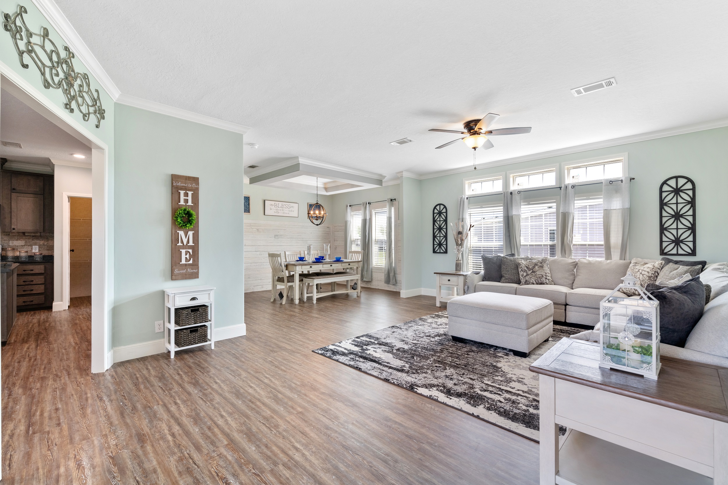 Spacious living room with light green walls, wood flooring, a gray sectional sofa, and a rug. Dining area with a wooden table is visible in the background. Airy and inviting.