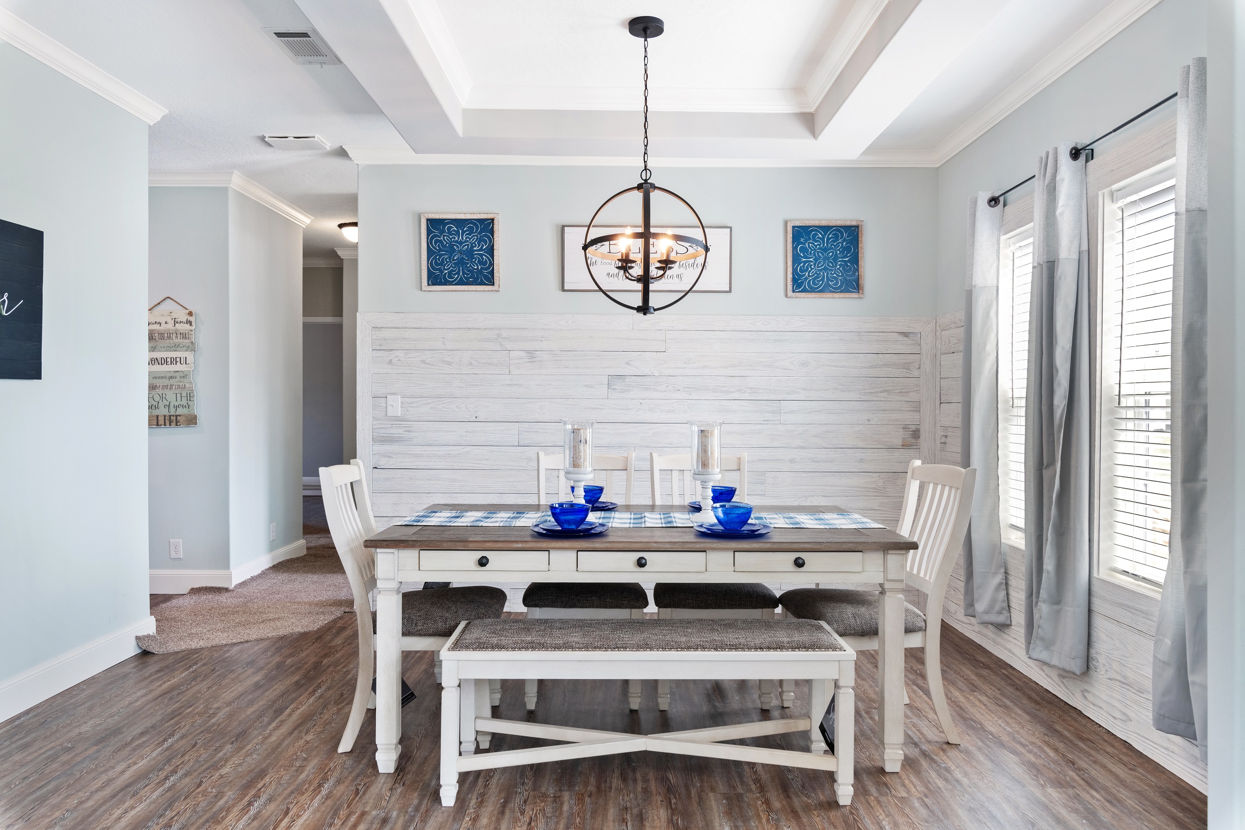 A bright dining room with a rustic table set, white chairs, and a bench. Blue accents on the table and artwork. Light wood flooring. Cozy and inviting.