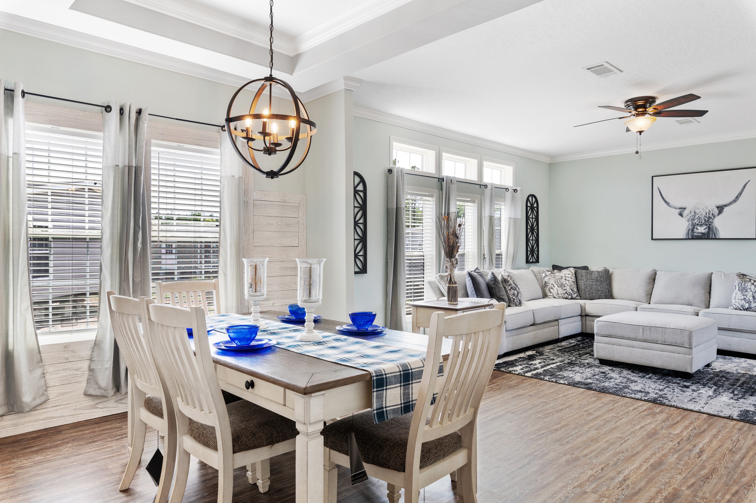 Bright, modern dining and living area with wood floors. Features a rustic table with blue dishes, a cozy gray sectional, and a bull artwork on the wall.