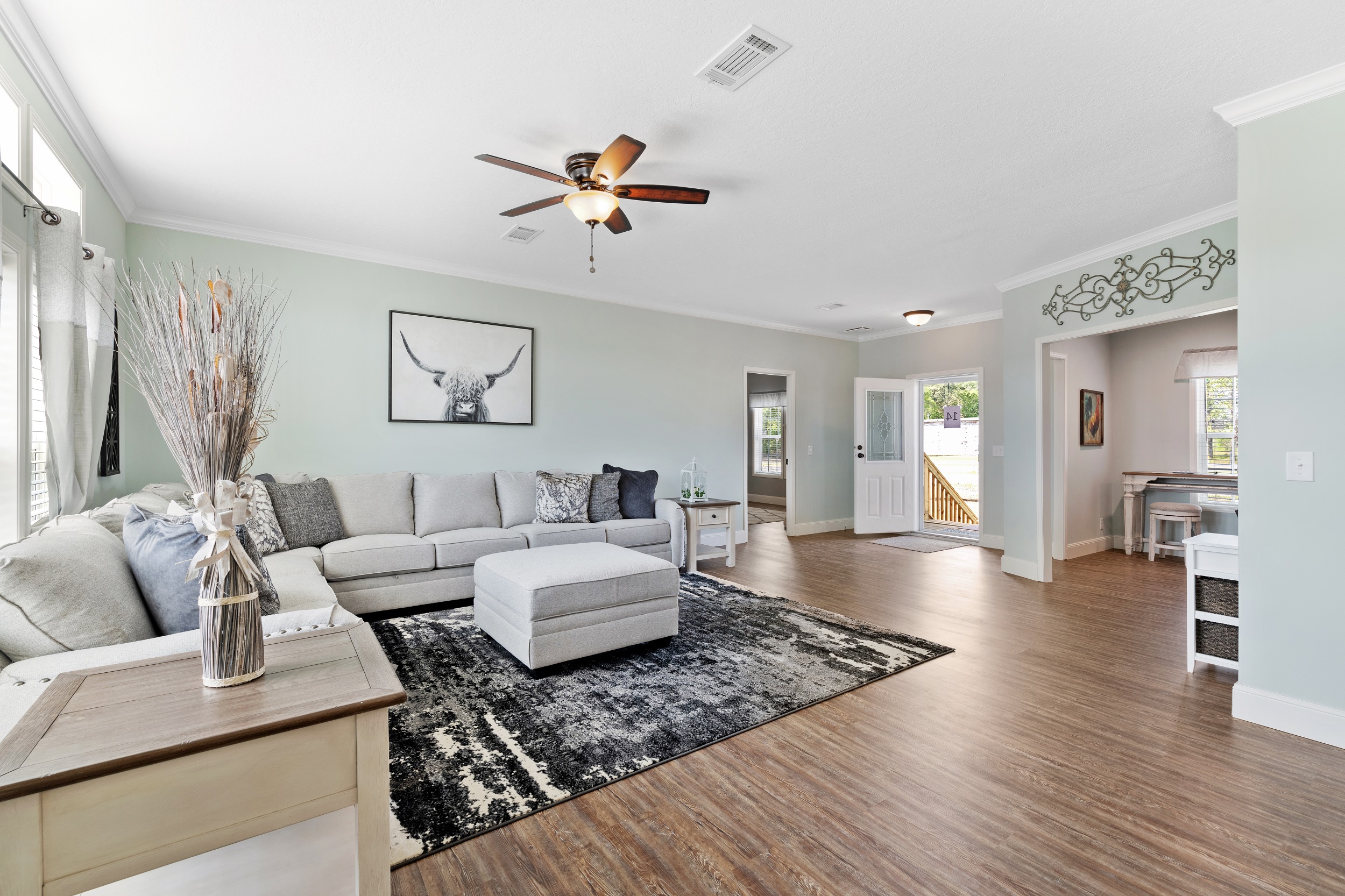 Spacious living room with light green walls, hardwood floor, gray sectional sofa, and black-and-white rug. A ceiling fan and a bull artwork add elegance.