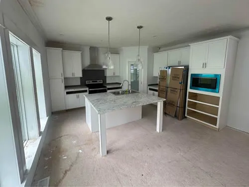 Modern kitchen with white cabinetry, a marble island, and stainless steel appliances. Large windows provide natural light. Cardboard boxes on the floor suggest recent renovations.
