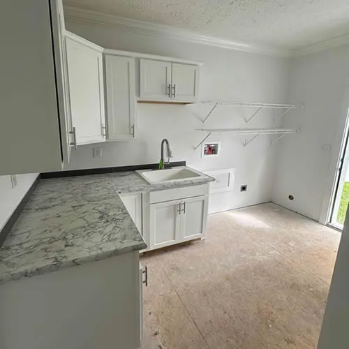 A bright, unfinished laundry room with white cabinets, a marble countertop, and a sink. Two wire shelves and empty flooring suggest ongoing renovation.