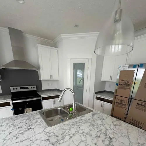 Modern kitchen with marble countertops, stainless steel appliances, and white cabinets. A large pendant light hangs above the central island. Cardboard boxes are stacked near the fridge.