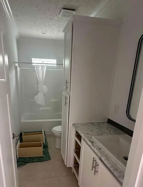 Narrow bathroom with a marble countertop, under-construction cabinets, and a shower. A tied curtain on the shower and exposed cabinet interiors.