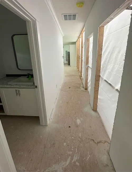 Long, dimly lit hallway under renovation with plastic-covered door frames and an unfinished floor. A small sink and mirror are visible in the left room.