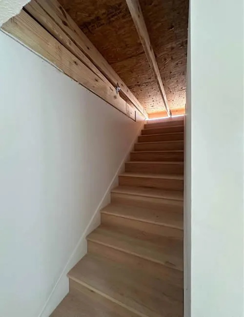 A narrow staircase with wooden steps, leading upwards to a low ceiling of exposed beams. Sunlight filters through a small gap, creating a cozy yet cramped atmosphere.