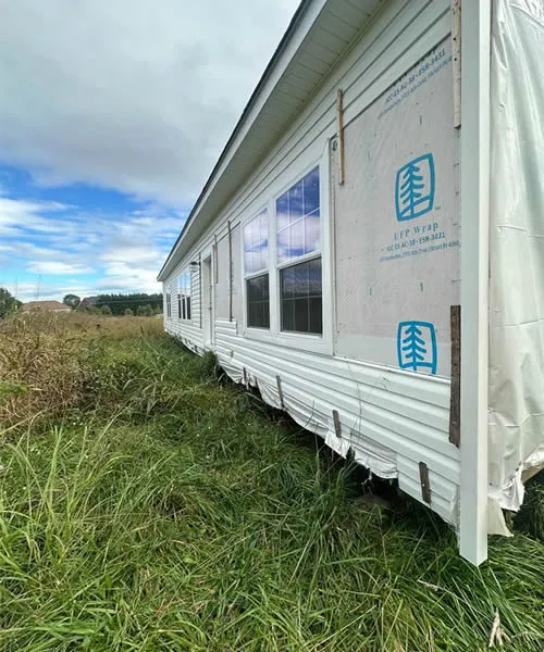 A partially wrapped, unpainted house stands in overgrown grass beneath a cloudy sky. Windows reflect the blue sky, creating a sense of abandonment.