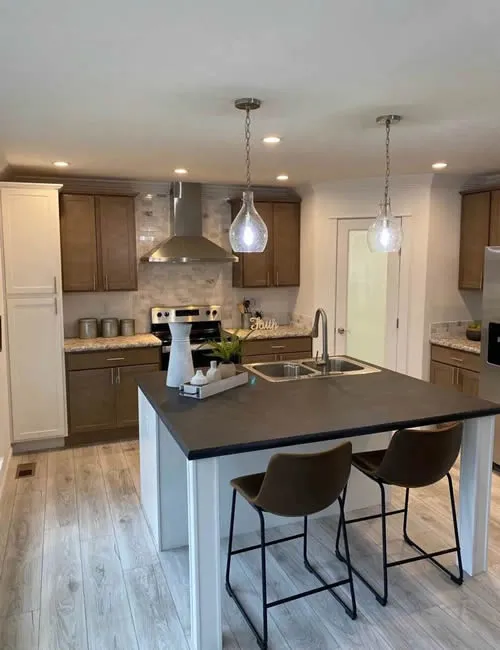 Modern kitchen with wooden cabinets, gray island, and pendant lights. Features include a stove, stainless steel hood, and bar stools. Bright and welcoming.