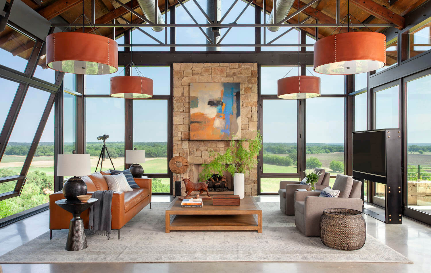 Spacious, modern living room with large windows, leather sofa, gray armchairs, stone wall, abstract art, and a view of green fields. Cozy, airy ambiance.