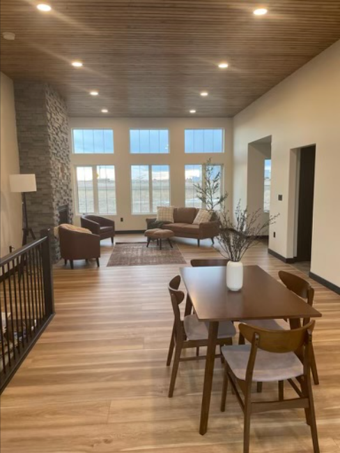 Spacious living room with wooden ceiling and floors, stone fireplace, and large windows. Modern furniture includes a wooden table and cozy chairs.