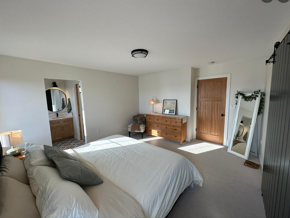 A cozy bedroom with a neatly made bed, light bedding, and gray pillows. A wooden dresser with a lamp and framed photo sits on the right. An open door leads to a bathroom. A full-length mirror rests against a wall, reflecting natural light streaming in from a window. The overall feel is calm and inviting.