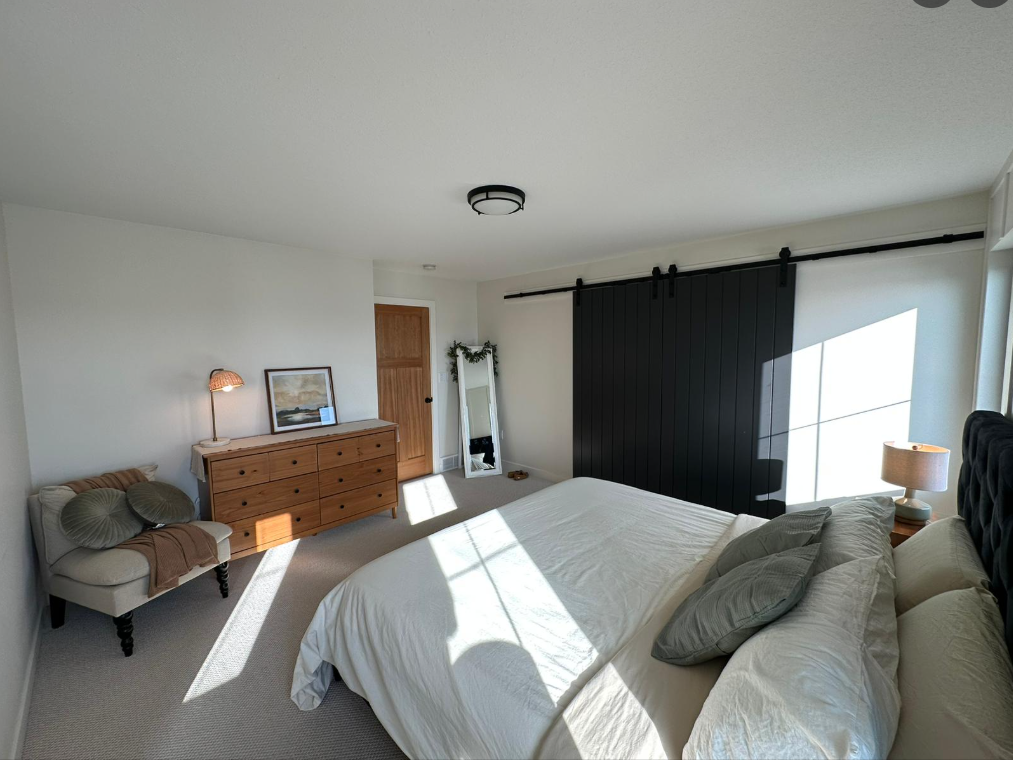 A bright bedroom with a large bed featuring white linens and gray pillows. A wooden dresser and round lamp sit to the left, beneath a painting. A black sliding barn door and a full-length mirror enhance the room's modern, cozy vibe.