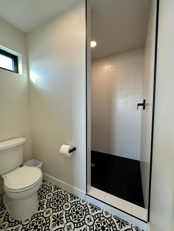 Bathroom with black-and-white patterned floor tiles, a white toilet, and a roll of toilet paper on the wall. A glass door reveals a tiled shower.