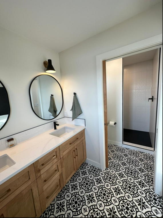 Stylish bathroom with patterned black-and-white tile floor, wood vanity, twin sinks, round mirrors, and a walk-in shower. Modern and clean design.
