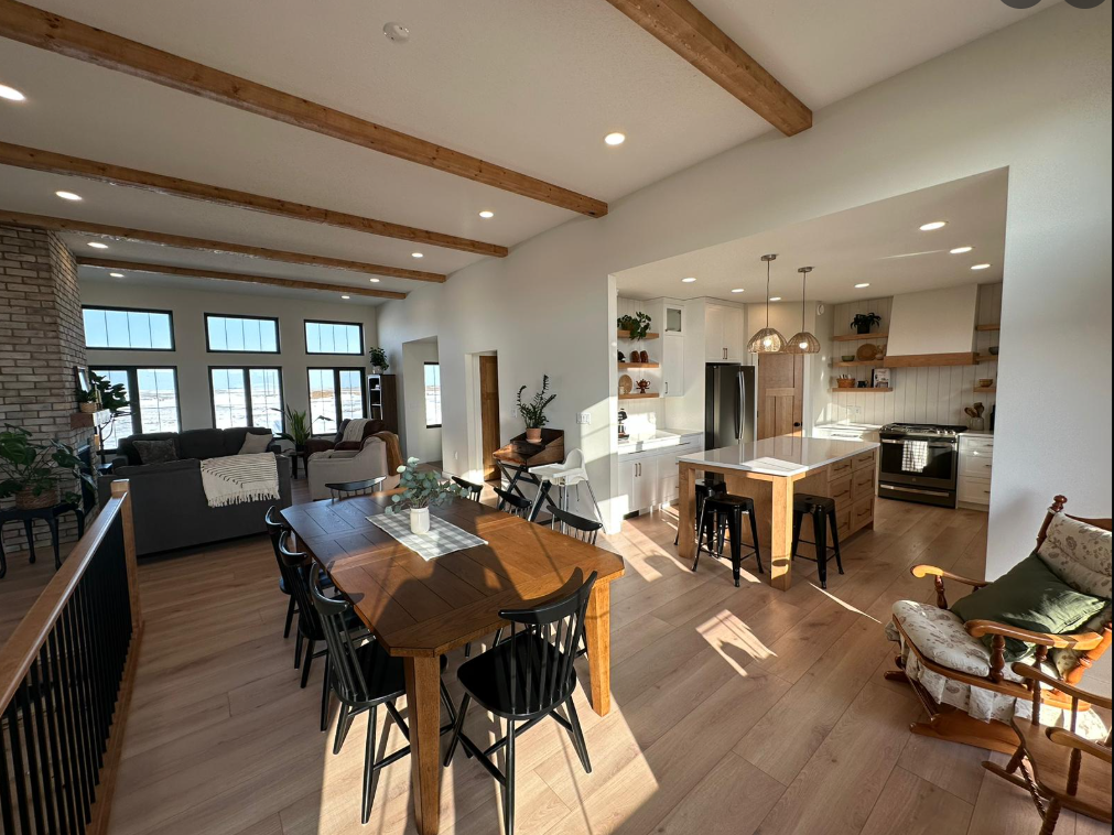 Open-concept living space with a modern kitchen, dining area, and cozy seating. Wood beams, abundant natural light, and neutral tones create warmth.