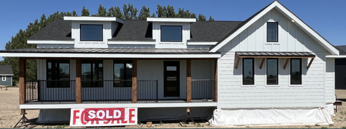 A modern, white house with a porch and black roof in a rural setting. A "Sold" sign covers a "For Sale" sign, indicating a recent purchase. Trees line the background.