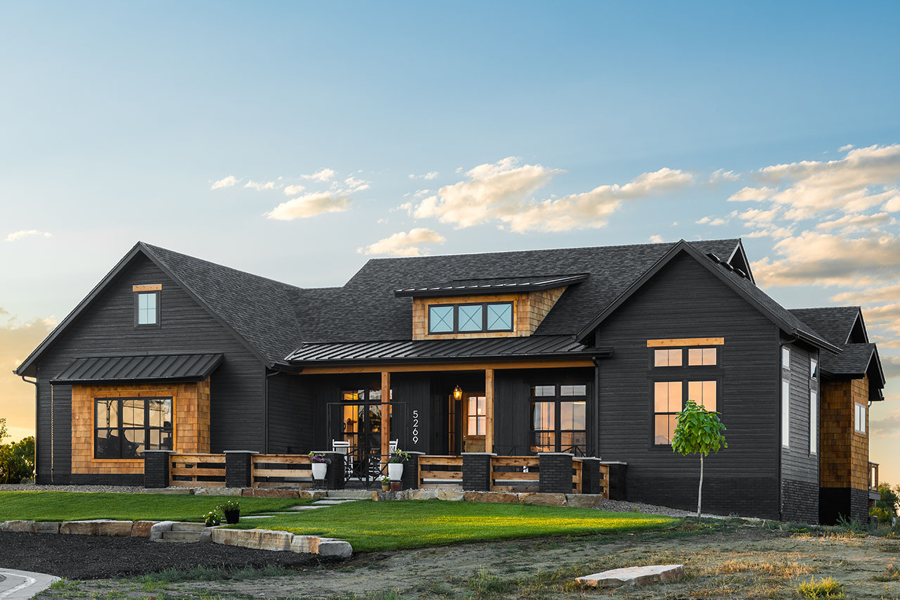 A modern black house with wood accents sits under a blue sky with scattered clouds. The front porch has seating, and a small tree is planted in the yard.