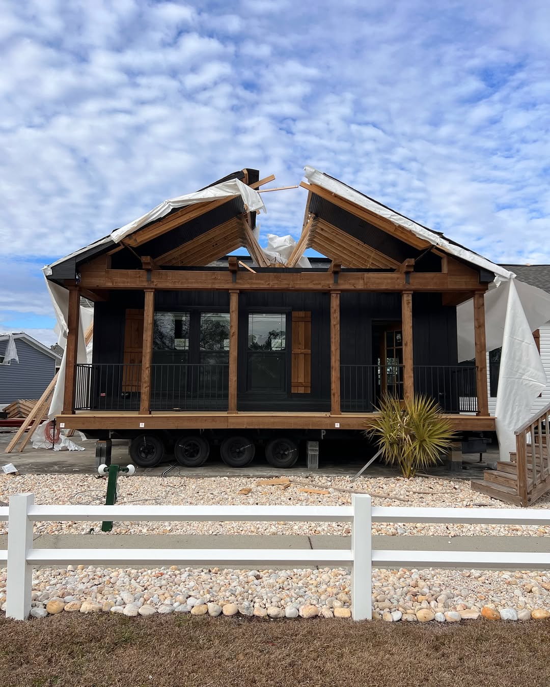 A tiny house on wheels with a black exterior and wooden accents sits on a gravel lot. Its roof is open, partially covered by white tarps. A white fence borders the front.