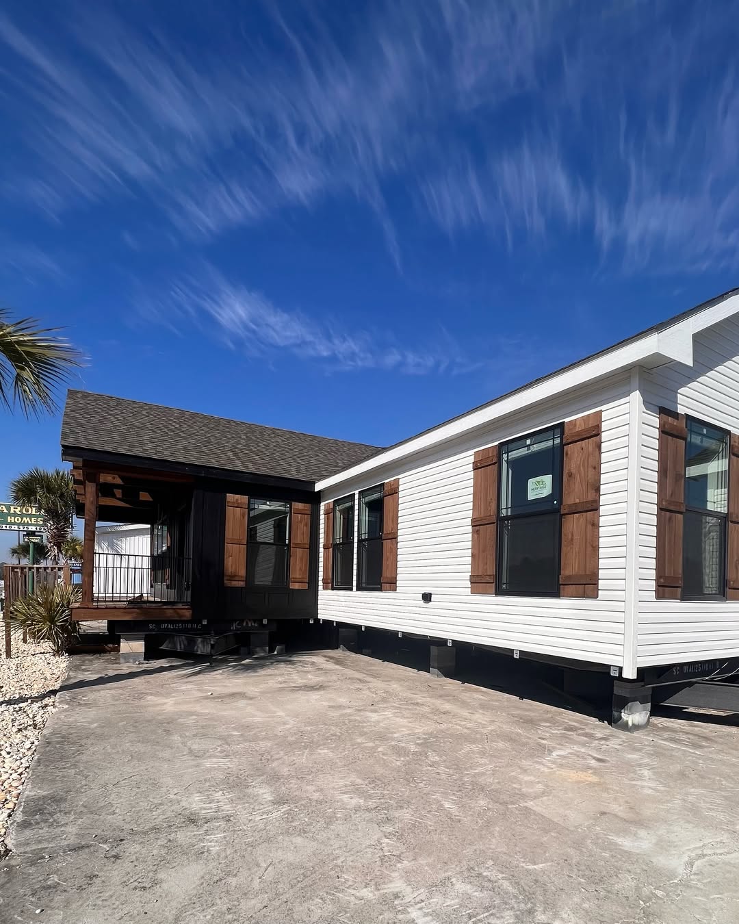 A modern white modular home with wooden shutters sits under a clear blue sky. The home is on a concrete lot, with palm trees visible, conveying a coastal feel.