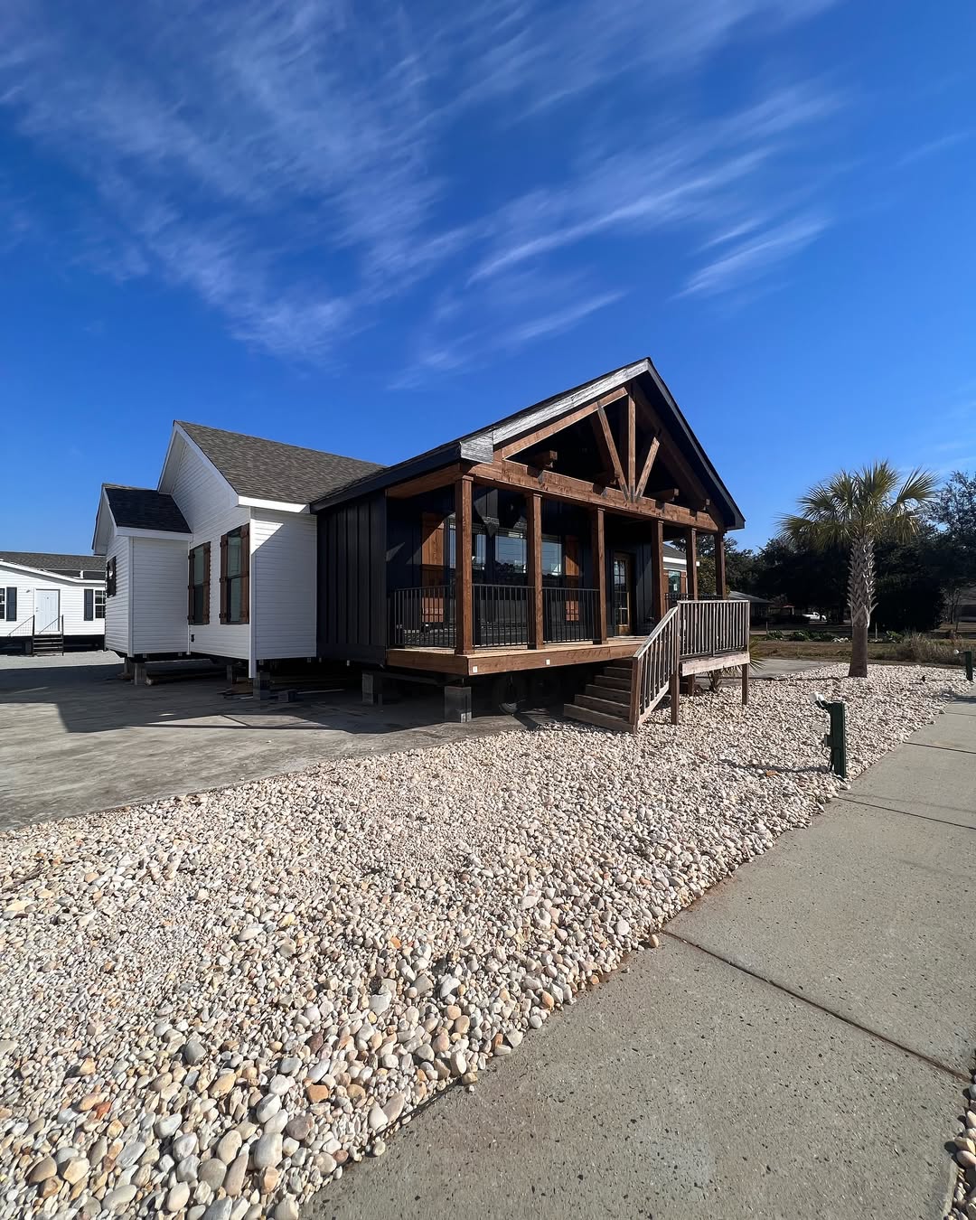 A modern house with a gabled roof and wooden porch against a blue sky. The foreground shows a rocky yard and sidewalk, creating a serene atmosphere.