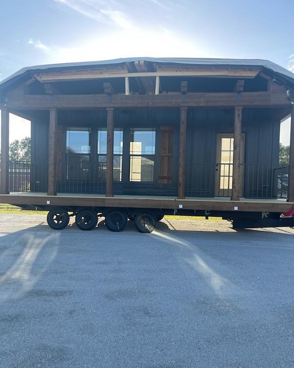 A small, dark wooden mobile cabin with large windows and a porch sits on a trailer. The bright sky casts a halo around the roof, creating a serene atmosphere.