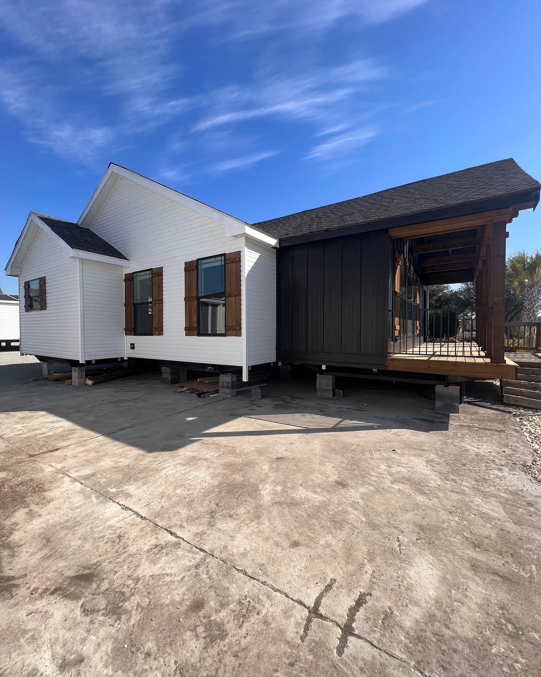 A white modular home with dark wood shutters sits elevated on blocks on a concrete lot. The blue sky above adds a serene atmosphere.