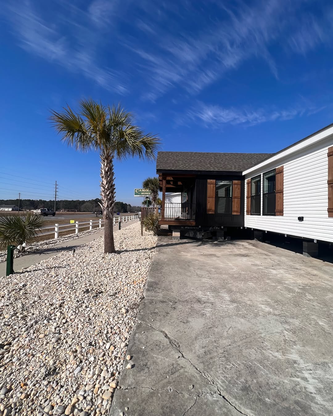 A manufactured home with wooden shutters sits on a gravel-lined lot with a palm tree. The sky is clear blue, creating a peaceful, sunny atmosphere.