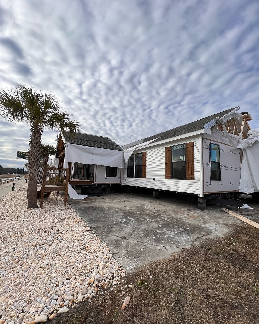 Damaged white house with a tarp-covered roof stands on a concrete lot. A palm tree is nearby under a dramatic sky with wavy clouds, creating a somber tone.