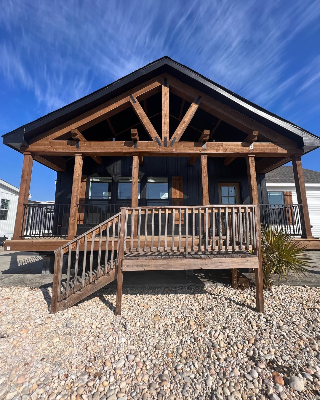 Wooden cabin with a gabled roof and front porch, set against a clear blue sky. The entrance is elevated, with wooden railings and surrounded by pebbles.