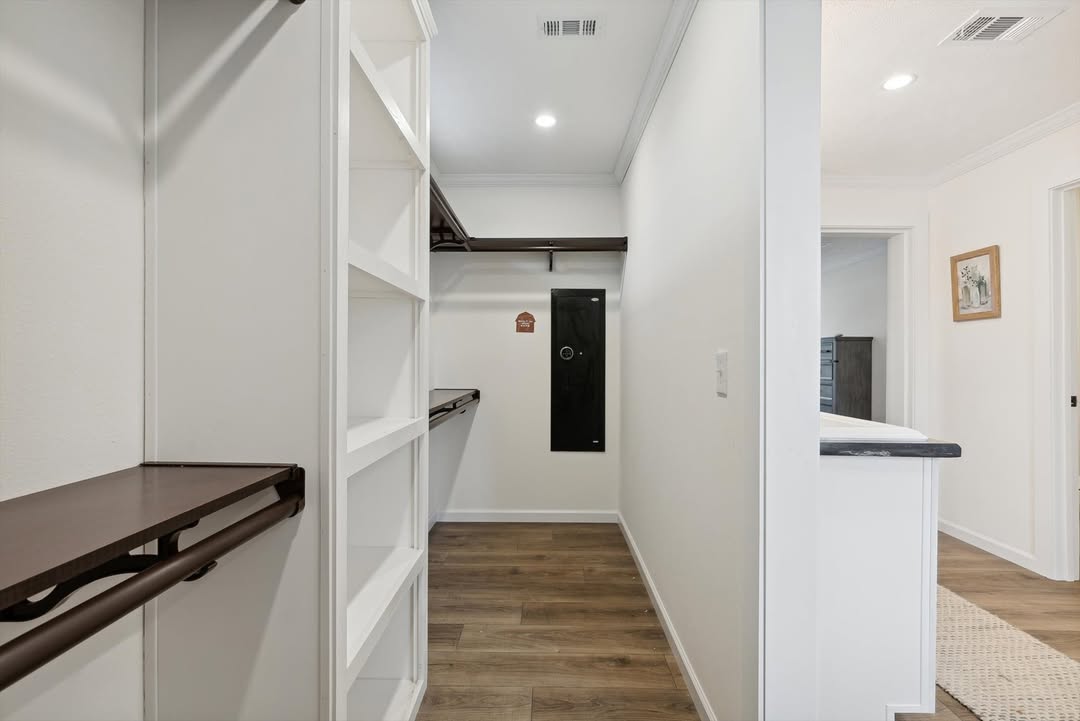 Spacious walk-in closet with white walls, wooden flooring, and dark shelves. Lit by ceiling lights, it connects to a room with a light-colored carpet.