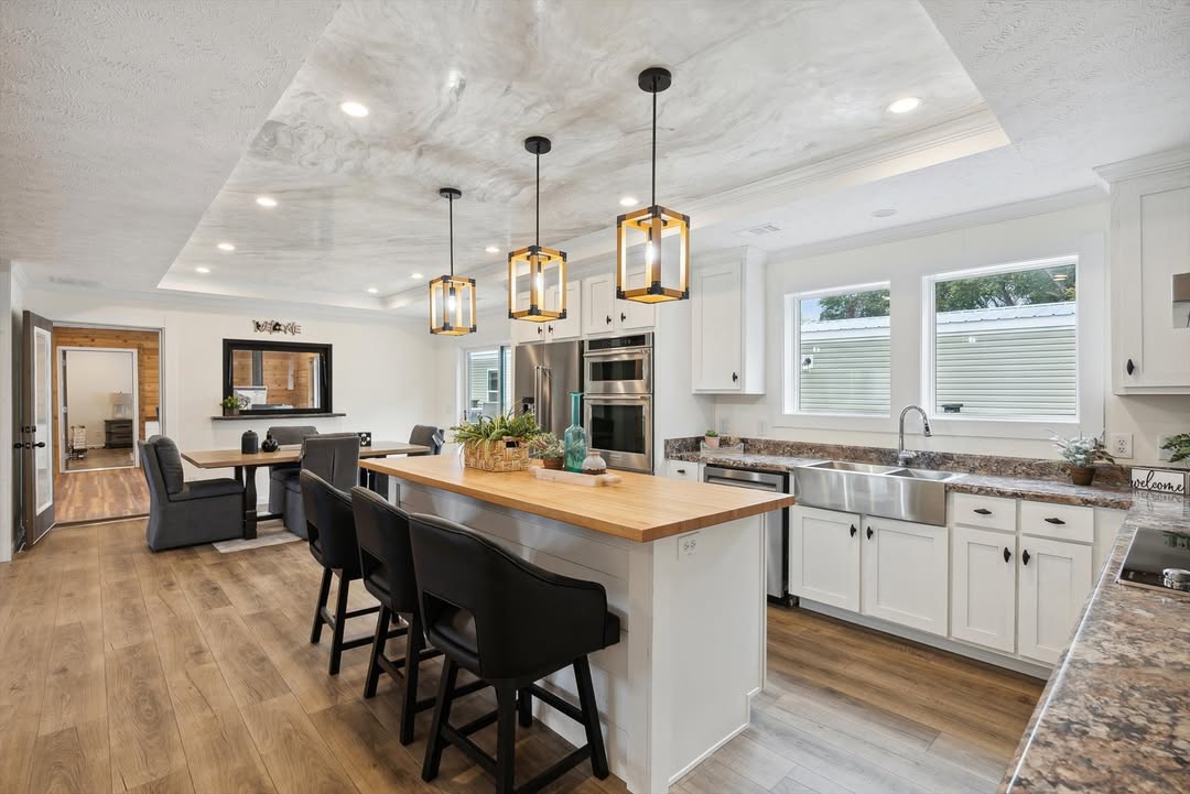 Spacious modern kitchen with a large island, three pendant lights, black bar stools, and wooden flooring. White cabinets and stainless steel appliances.